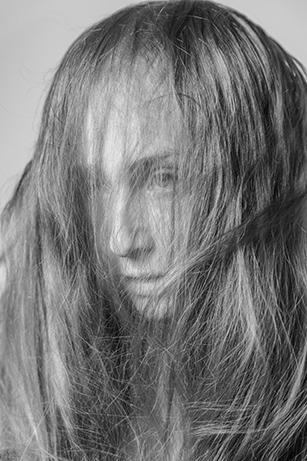 A black-and-white close-up portrait of a person with long hair partially obscuring their face, looking towards the camera.