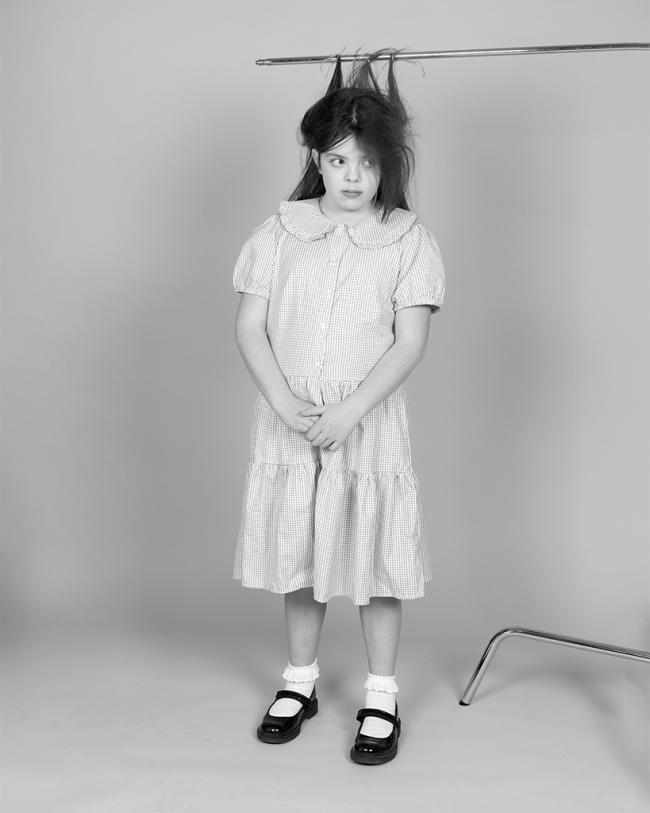 A young child stands in a dress beneath a clothing rail, with sections of hair tied upward to the bar in a surreal studio portrait.