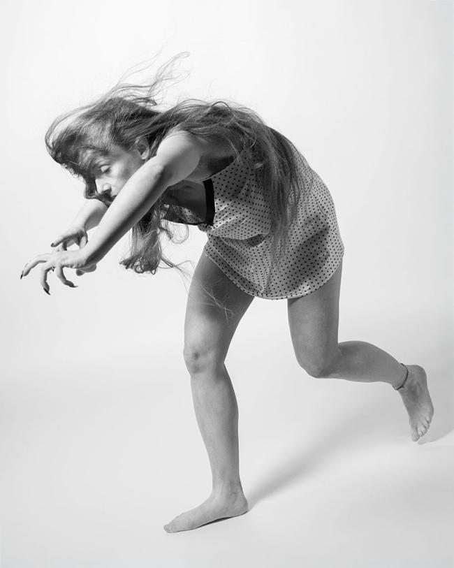 A person in motion bends forward with arms outstretched in a dynamic black-and-white portrait, hair sweeping through the frame.