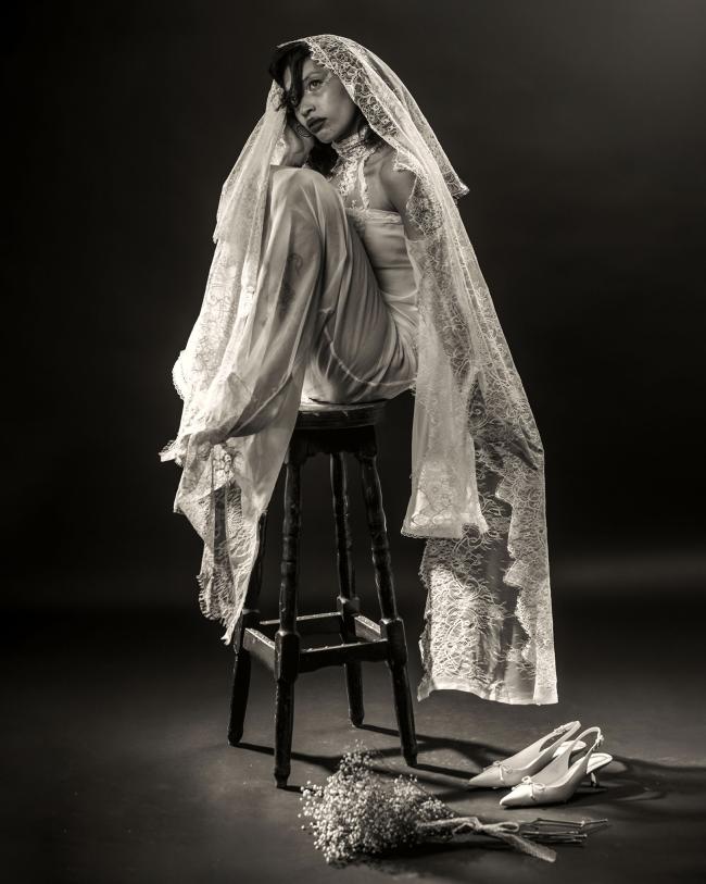 Third model sitting on a chair wearing a long white silk bridal dress against a black backdrop