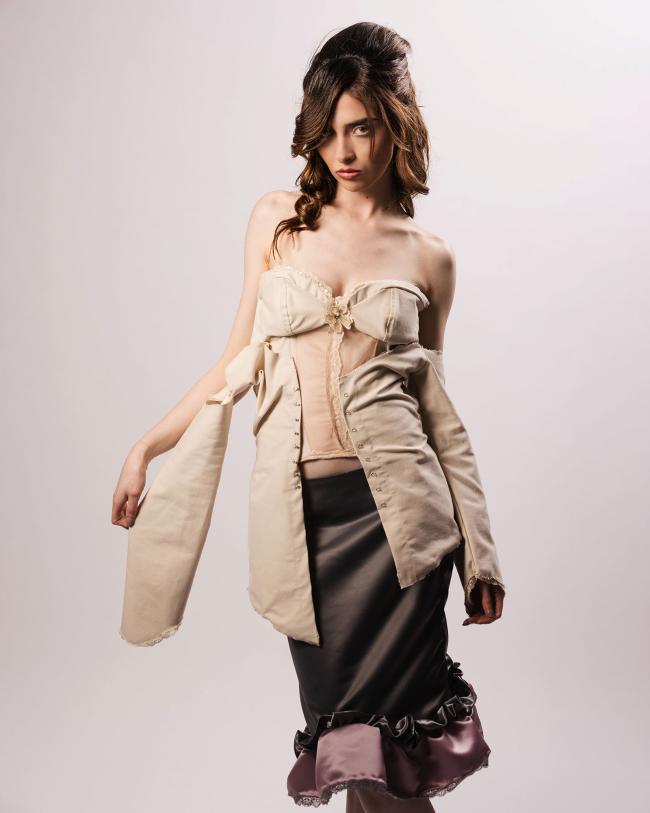 First model wearing a grey satin skirt and beige draped corset, holding one sleeve of the garment in her hand, standing against a white backdrop
