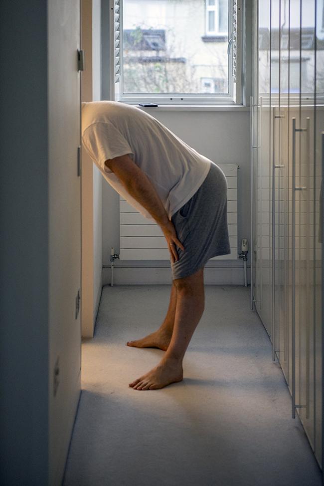 A person bends forward in a bright room, partially hidden behind a doorway in a quiet domestic scene.