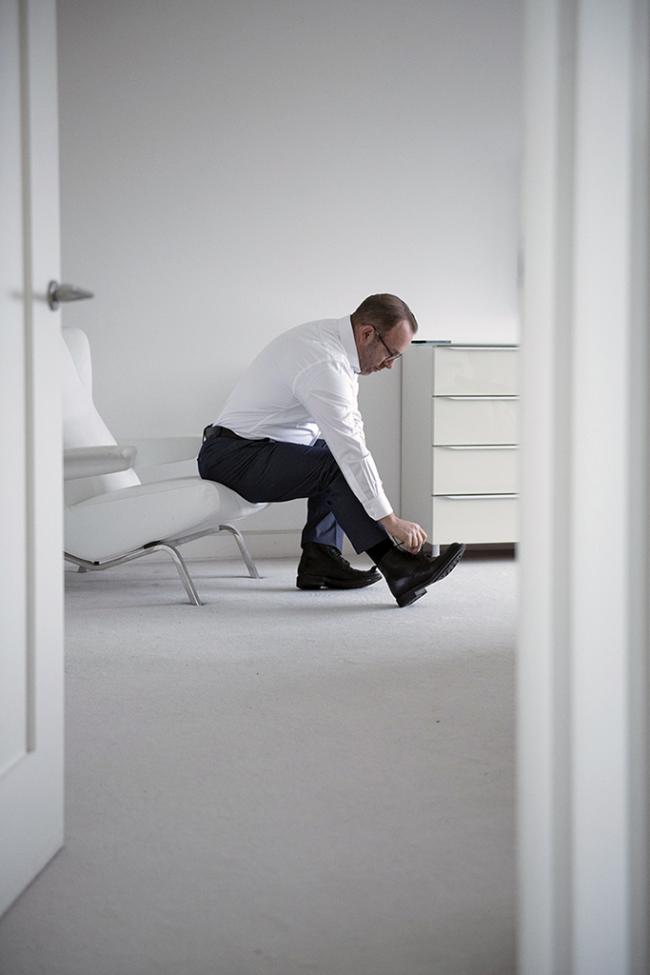 A person in formal clothes sits on a chair fastening a shoe in a bright minimalist room.