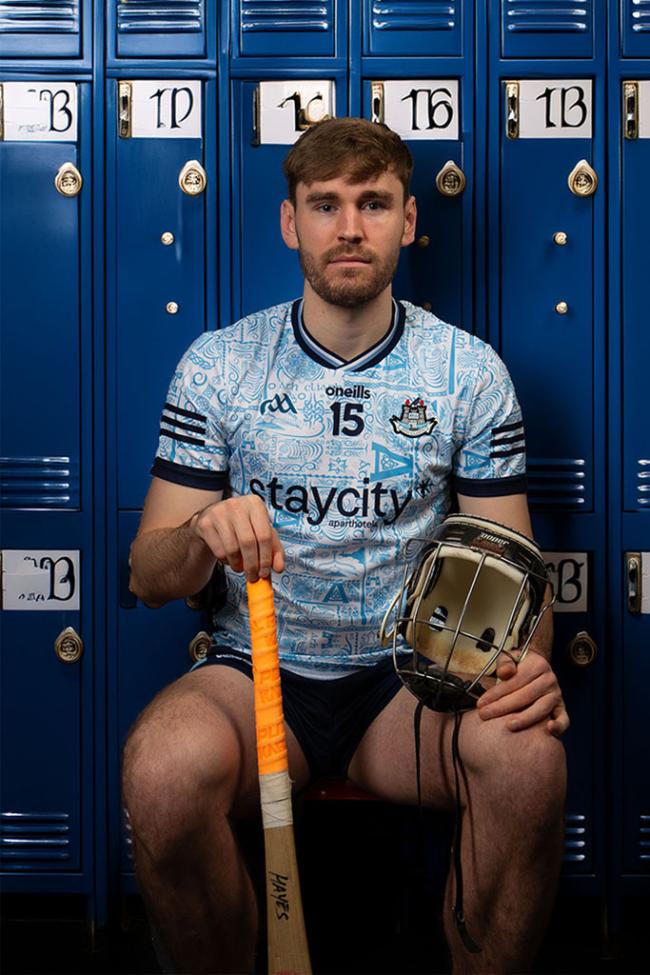 A hurler in a Dublin jersey sits in front of blue lockers holding a helmet and resting a hurl on the floor. 