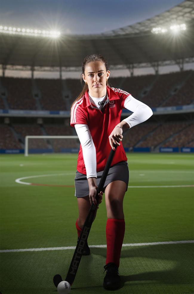 A hockey player in a red uniform crouches on a floodlit pitch, holding a hockey stick with a ball at her feet.