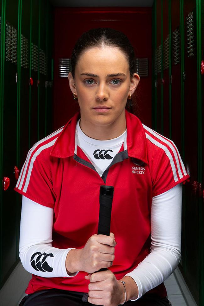 A hockey player in a red uniform sits in front of lockers, holding a hockey stick and looking directly at the camera.
