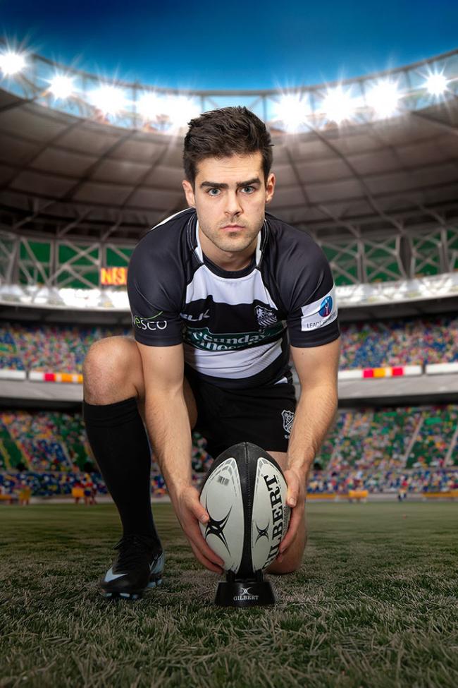 A rugby player in a black and white kit kneels on a pitch, positioning a rugby ball under bright stadium lights.