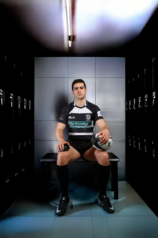 A rugby player in a black and white kit sits holding a rugby ball in a dark locker room.