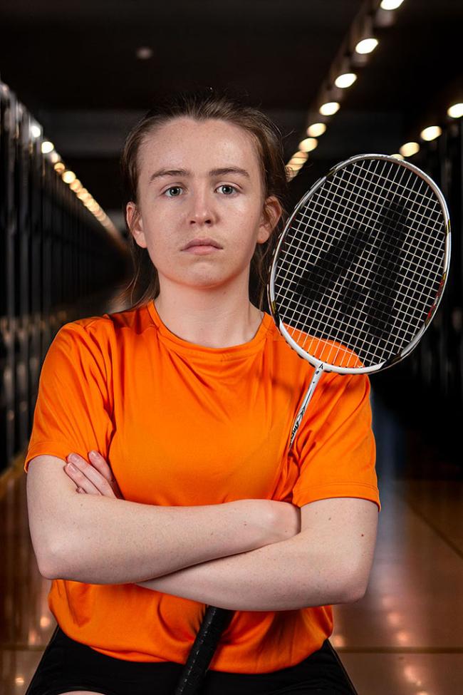 A badminton player in an orange shirt stands with folded arms, holding a racket in an indoor sports hall.
